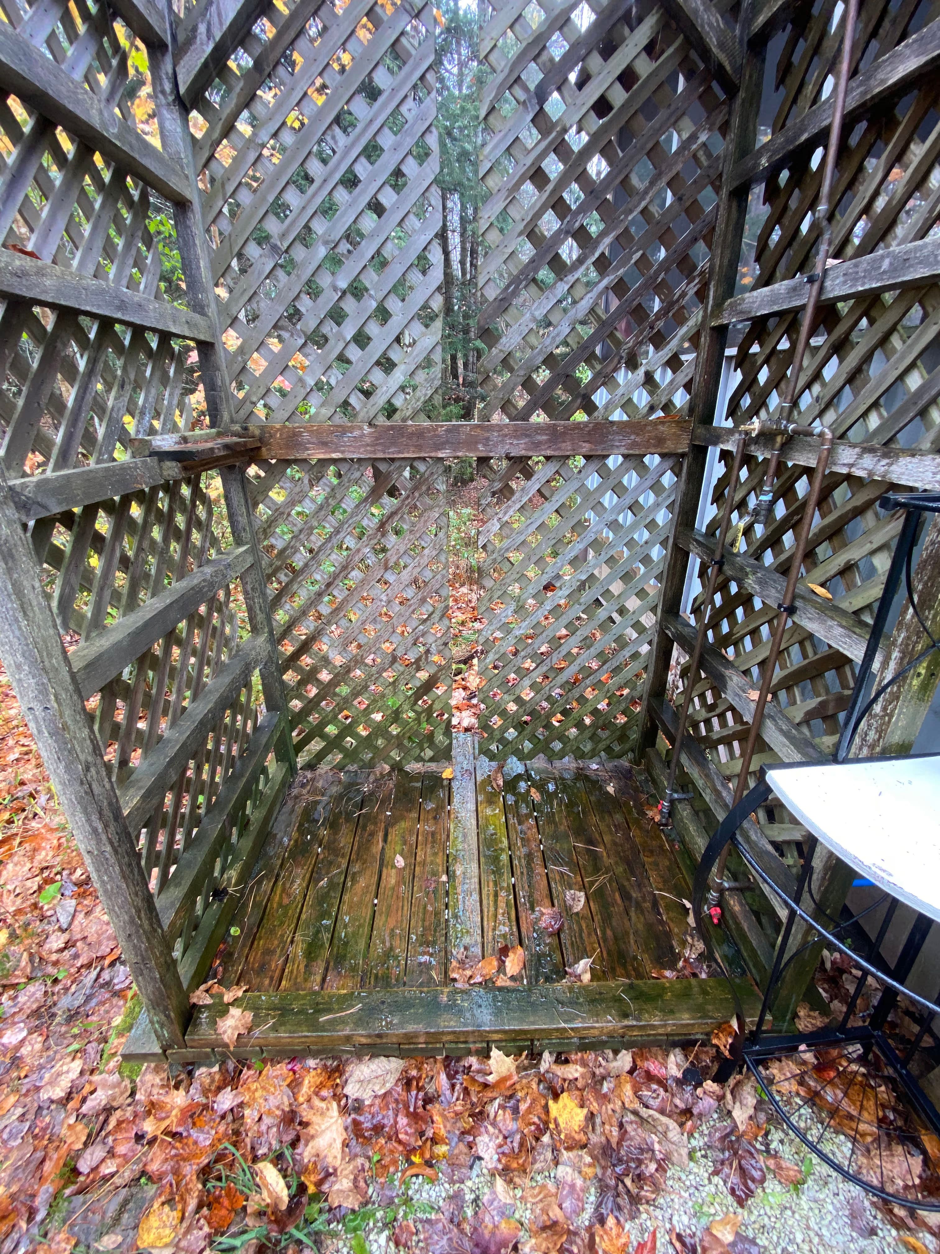 Empty outdoor shower stall with wooden lattice walls, wet floor, and fallen leaves.