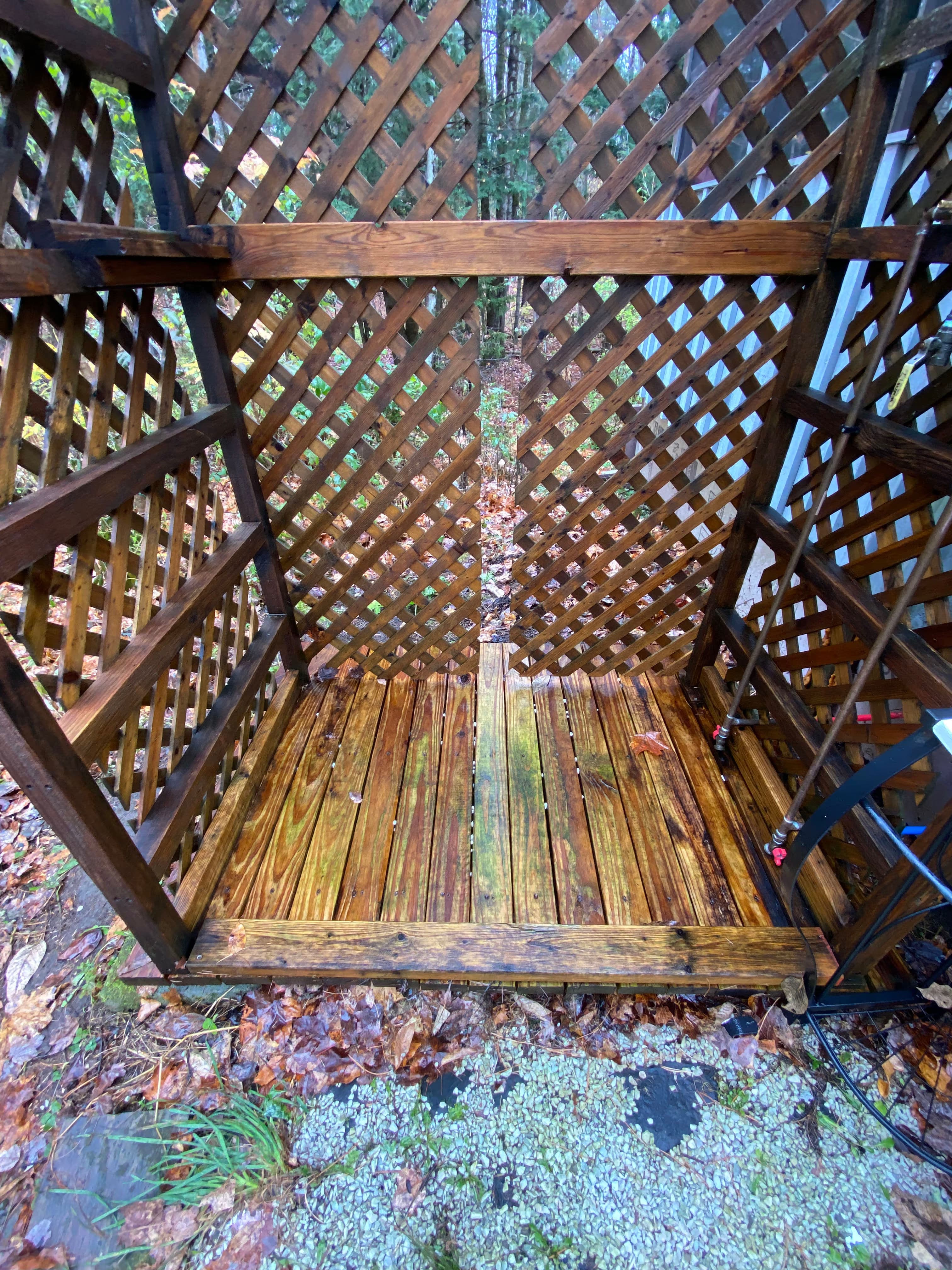 Outdoor wooden shower enclosed by lattice, featuring a wet wood floor and natural surroundings.